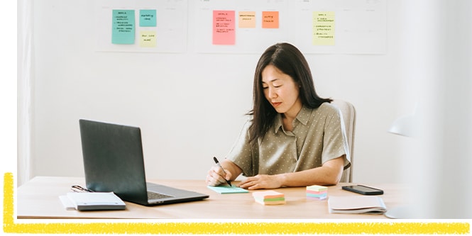 Une femme assise à son bureau, dressant une liste de tâches à accomplir et utilisant des notes Post-it®
