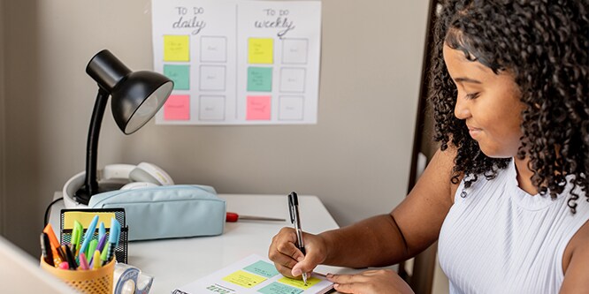 Étudiant prenant des notes sur des notes Post-it®, avec en arrière-plan un planning de révisions détaillant les tâches quotidiennes et hebdomadaires.
